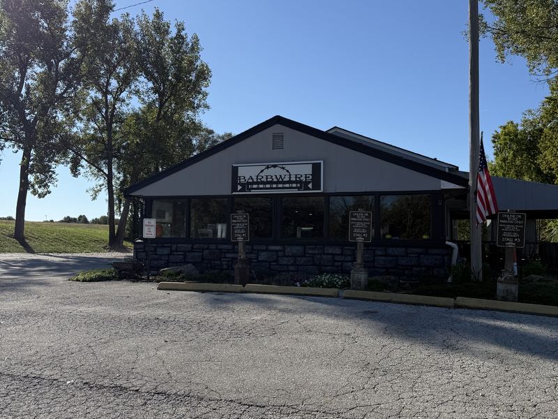 Barbwire Barbecue on a sunny day: a gray building with a stone front, its sign over the door and an American flag out front, with tall trees behind