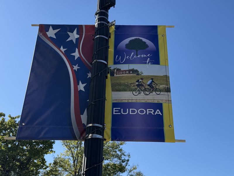 Two banners on a downtown streetlight against a clear blue sky: a stars-and-stripes banner and a Welcome to Eudora banner with bike riders