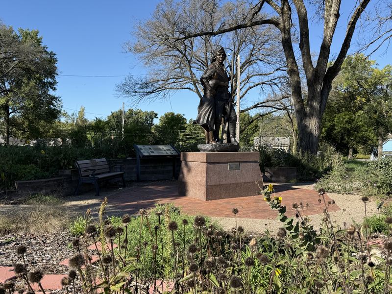 The bronze statue of Shawnee leader Paschal Fish and his daughter Eudora on a brick base, in a garden surrounded by trees on a sunny day