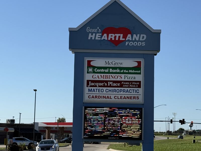 The tall blue Gene's Heartland Foods sign with a red heart, listing the shops in the same center, against a clear blue sky